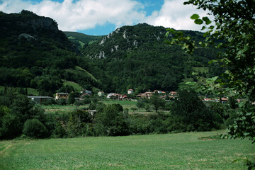 Scenic landscape of a green meadow, small traditional houses of a Spanish village, and forest-covered mountains. Blue sky with soft white clouds. Typical rural scenery of Asturias