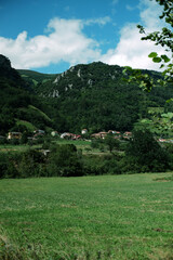 Scenic landscape of a green meadow, small traditional houses of a Spanish village, and forest-covered mountains. Blue sky with soft white clouds. Typical rural scenery of Asturias