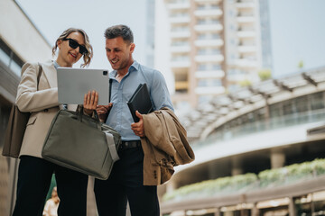 A couple in business attire stands outdoors in a modern city, smiling as they view a tablet. They carry bags and jackets, suggesting a day of collaboration and urban work.