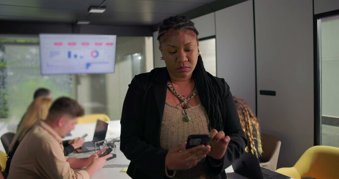 Focused African American businesswoman using smartphone during office meeting, team working in background, data analytics screen visible on wall - Powered by Adobe