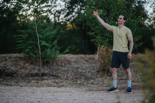 A fit man in a yellow shirt and dark shorts stands on a dirt path in a wooded area, raising his arm as if signaling or greeting, with a backpack nearby. - Powered by Adobe