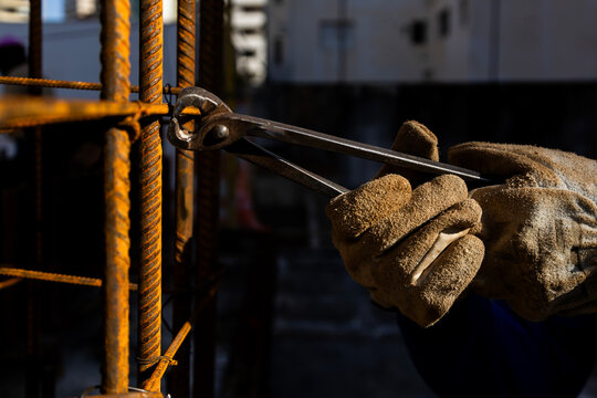 Close-up of a construction worker wearing protective gloves and using pliers to cut or tie steel rebar at a job site. Warm light highlights the texture of rusted metal and manual labor.