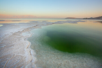 Low-angle view of the Dead Sea shore near Ein Bokek, Israel, showing massive white salt crystal formations and the perfect reflection of the golden Judean Desert mountains and resort hotels