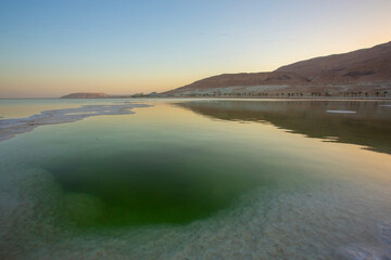 Low-angle view of the Dead Sea shore near Ein Bokek, Israel, showing massive white salt crystal formations and the perfect reflection of the golden Judean Desert mountains and resort hotels