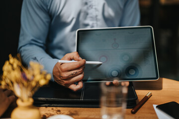 A professional uses a tablet and stylus to review charts on screen during a focused business meeting at a wooden desk, with a plant and coffee nearby for a natural workspace feel.