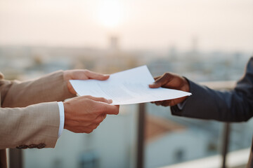 Two professionals exchanging a contract on a rooftop terrace at golden hour — close-up of hands passing important business paperwork and agreement