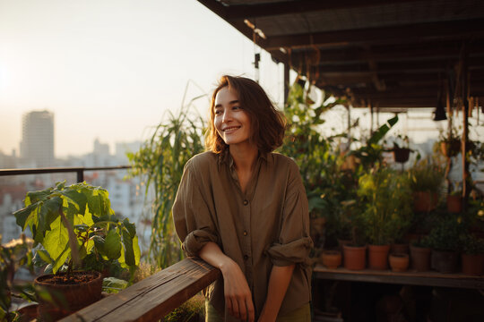 Smiling young woman relaxing on a plant-filled rooftop balcony at golden hour, enjoying urban gardening, warm sunlight and a serene city escape - Powered by Adobe