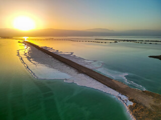 High-angle aerial view of geological sinkholes (collapse pits) forming along the rapidly receding shoreline of the Dead Sea. The image features contrasting colors of water-filled holes 