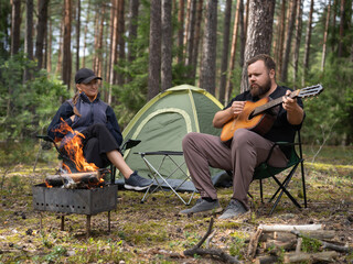 Bearded man playing guitar for his partner, waiting for coals for barbecue and sitting near a tent in the forest while hiking, enjoying the calm atmosphere and fresh air