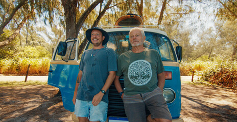 Two relaxed men stand near the retro bus loaded with surfing boards and watch somewhere. Modern hippies travel by retro bus and check waves on a surfing spot