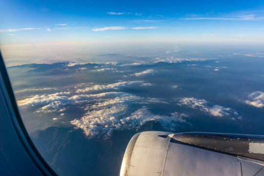 Panoramic aerial view of layered mountains and clouds from high altitude airplane window showing engine cowling and brilliant sunlight reflection.
