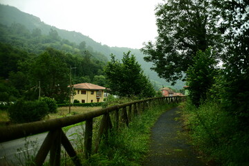 Scenic walking trail with a wooden fence leading into the distance, surrounded by countryside with lush vegetation, traditional houses, and forested mountains. Senda verde. Hiking in Asturias