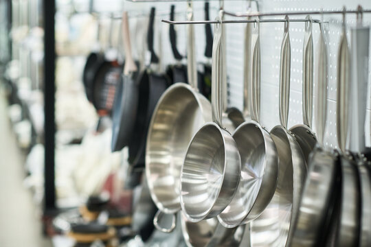Stainless steel frying pans and saucepans hanging on hooks in kitchenware store, cookware displayed in organized row with blurred background showing additional utensils and equipment