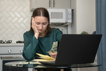 Young woman looking stressed while managing her home finances using her laptop and counting cash, highlighting financial struggles and budgeting challenges