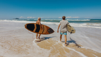 Young and elderly surfers walk with surfing boards on the beach in Brazil