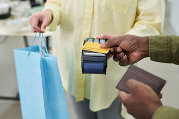 Caucasian woman handing shopping bag while Black man making payment with credit card using point of sale terminal, both adults engaged in retail transaction, close up on hands