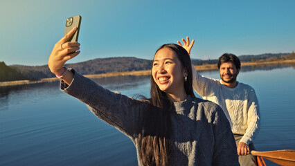 A young Asian woman smiles in a selfie, a man waves in the background. They canoe on a serene lake at sunset, enjoying a joyful outdoor adventure and bonding moment.