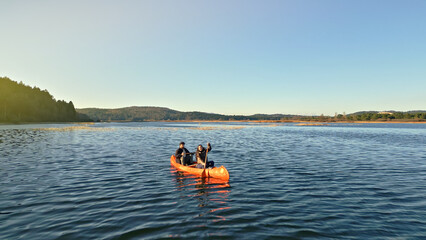 A couple paddles an orange kayak on a serene lake, surrounded by rolling hills. The calm water reflects the clear blue sky, capturing a moment of tranquility and connection in nature.