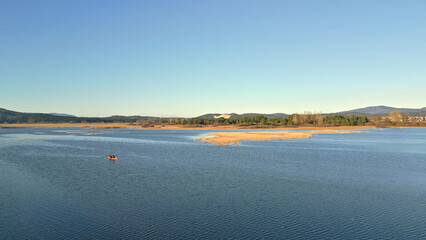 A high-angle view of a couple paddling a canoe on a serene lake, surrounded by distant mountains and a clear blue sky. The tranquil setting highlights weekend activities and an active lifestyle.