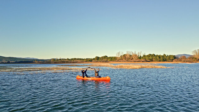 A couple paddles an orange kayak on a serene lake, surrounded by autumnal trees and distant mountains. The clear blue sky and calm waters create a peaceful setting. - Powered by Adobe
