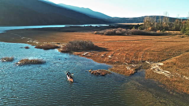 A couple paddles a canoe on a serene river, surrounded by autumnal fields and distant mountains. The scene captures a peaceful weekend activity, emphasizing connection. - Powered by Adobe
