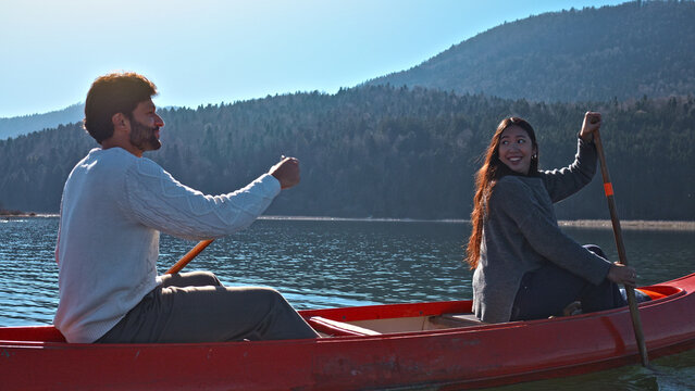A couple paddles a red canoe on a serene lake, surrounded by forested mountains. The man, in a white sweater, and the woman, in a gray coat, both smile, enjoying their weekend adventure. - Powered by Adobe