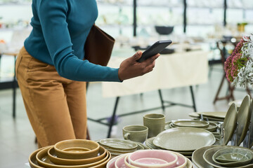 Young adult Black woman holding smartphone while standing near ceramic tableware display in store,...