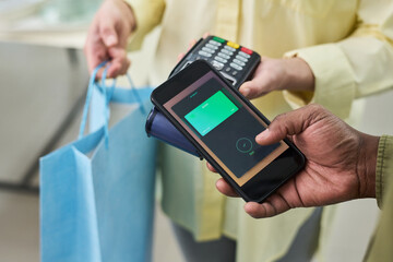 Black young adult man holding smartphone making contactless payment while Caucasian woman holding shopping bag and payment terminal during retail transaction