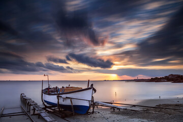 Long-exposure coastal scene at sunset featuring an old wooden fishing boat resting on a sandy shore beside wooden docks. Ravda, Burgas, Bulgaria