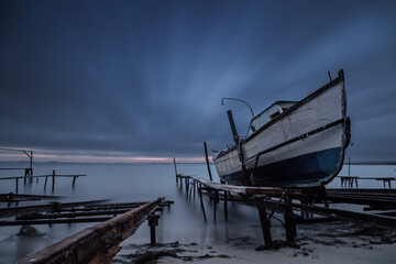 Long-exposure coastal scene at sunset featuring an old wooden fishing boat resting on a sandy shore beside wooden docks. Ravda, Burgas, Bulgaria