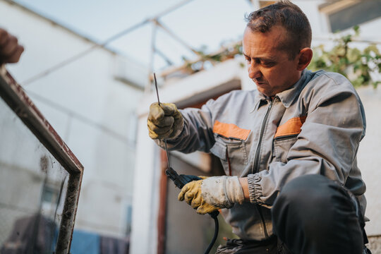 A focused male tradesman wearing a gray work suit with orange accents handles a tool to repair a glass panel at an outdoor construction site. Gloves protect hands during precision work.