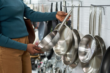 Unknown Black woman selecting stainless steel frying pan from display rack in kitchenware store,...