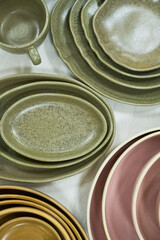 Stacked ceramic plates and bowls arranged on light surface, showcasing variety of shapes and muted earth tones, highlighting tableware assortment for kitchen or dining use