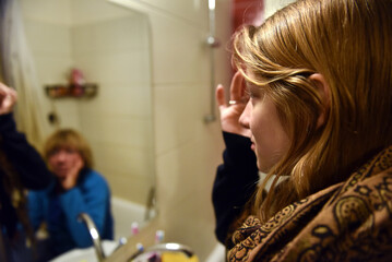 Young adult daughter applying face cream in a bathroom mirror with mother in the background