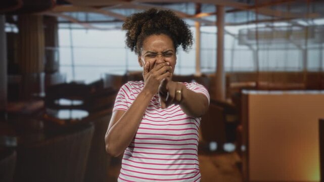 Woman young african american pointing finger and covering mouth while laughing in restaurant building wearing striped white pink shirt and curly hair pulled up; playful amusement.