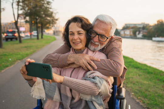 Senior couple woman in wheelchair caregiver old man making selfie enjoying walking. Elderly family man supporting embracing woman in chair for people with disability taking photo. Rehabilitation - Powered by Adobe
