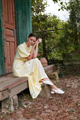 Woman in a yellow dress sits contemplative on rustic steps beside a doorway in an outdoor garden, exuding calm, thoughtful mood and serene rustic charm.