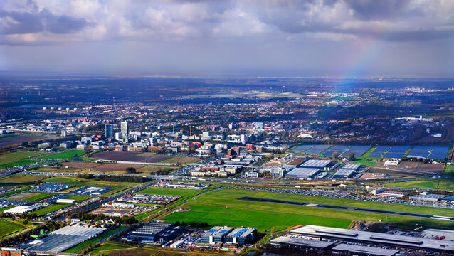 Aerial view of the landscape around Hoofddorp and Schiphol Airport in the Netherlands. A rainbow arches over the green fields, runways, and buildings under a dramatic cloudy sky