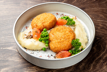 Crispy breaded fish or chicken cutlets served on a bed of creamy mashed potatoes with green peas, mushrooms, and fresh tomato garnish in a white bowl