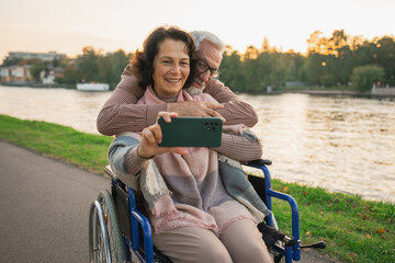 Senior couple woman in wheelchair caregiver old man making selfie enjoying walking. Elderly family man supporting embracing woman in chair for people with disability taking photo. Rehabilitation