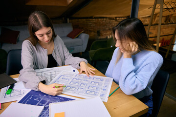 Two young women analyzing architectural drawings during project session. Concept of teamwork, professional research, planning workflow, and collaborative design development.