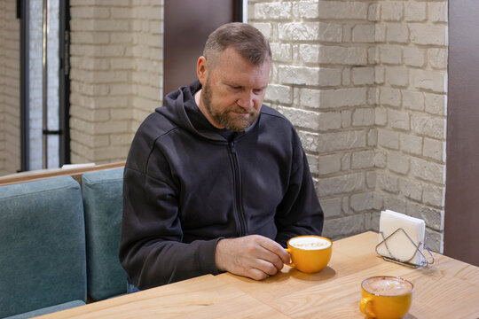 A middle-aged european man with beard in a dark hoodie sits in cozy cafe, looking thoughtfully at his orange cup of latte. Enjoying a quiet moment and coffee break.