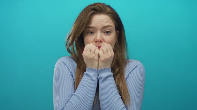Young caucasian woman with long brown hair holds clenched fists to mouth in studio setting with turquoise backdrop; anxiety.