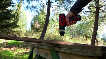 Close up footage of a man  using an electric cordless drill outdoors to make holes to a wooden plank.