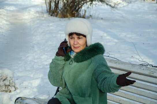 Senior woman sitting on snowy park bench, talking emotionally on smartphone with hand gesturing