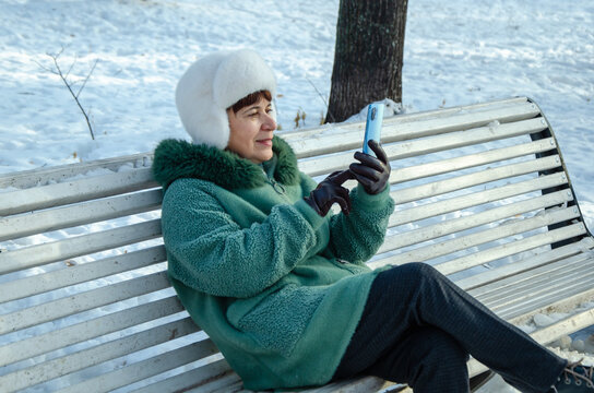 Senior woman in winter coat and white hat reclining on snowy park bench, smiling at smartphone screen