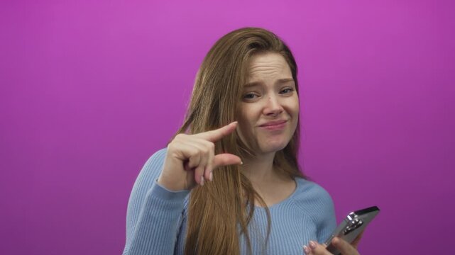 Young caucasian woman holding smartphone pinches fingers to show small amount in purple studio; doubt measurement size minimalism.