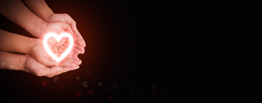 Bright neon heart resting in overlapping hands representing love and care, health care, CSR, insurance, family, World Heart Day, World Health Day, love, rescue, wellbeing, hope, support, compassion