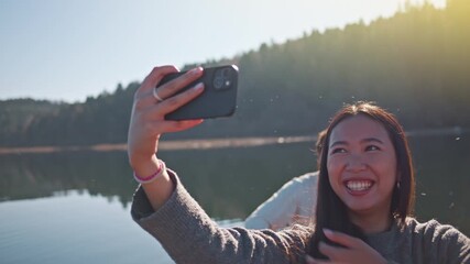 A young woman in a cozy sweater takes a selfie by a serene lake, arms raised, smiling joyfully. The sunlit backdrop of trees and water suggests a peaceful weekend getaway.