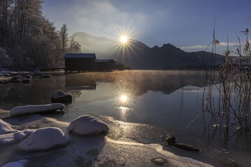 Evening mood at mountain lake in front of mountains, boat huts, shore, winter, snow, reflection, sunbeams, backlight, Lake Kochel, Alpine foothills, Bavaria, Germany